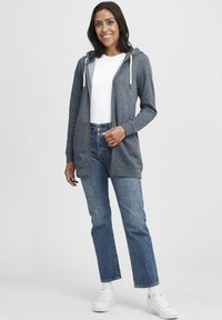 Grå zip-up hoodie med vita snören, långa ärmar och bruna accenter. Kombinerad med ljusblå raka jeans och vita sneakers.