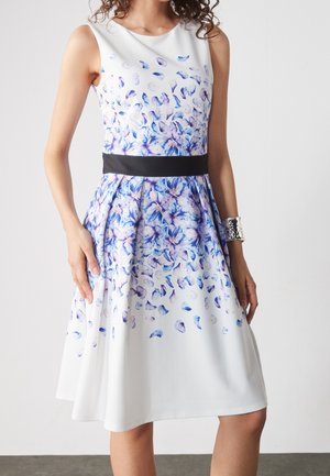 Ærmeløs hvid kjole med blå og lilla blomstermønster, sort taljebælte og sølvfarvet manchetarmbånd båret af en stående person.