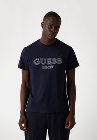 Navyblauwe katoenen t-shirt met een lichtgrijze "GUESS" logo, ronde hals, korte mouwen en een relaxte pasvorm.