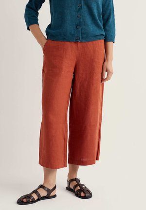 Woman wearing cropped burnt orange linen pants, teal buttoned cardigan, and black strappy flat sandals, standing with one hand in pocket.