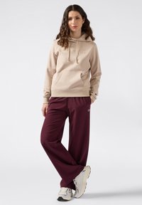 Felpa beige con cappuccio, dotata di tasca frontale e logo ricamato, abbinata a pantaloni bordeaux comodi e sneakers bianche.