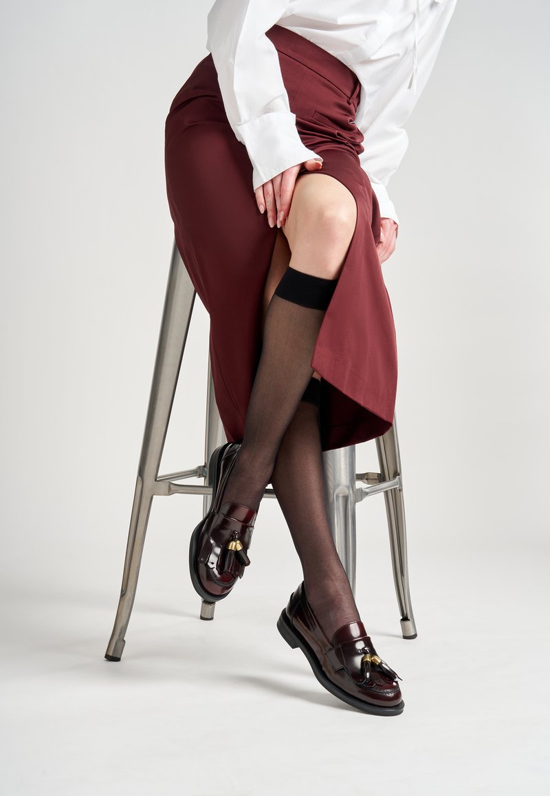 Mocasines de cuero burdeos con detalles de borlas, combinados con medias negras transparentes y una falda color burdeos, sentada en un taburete de metal.