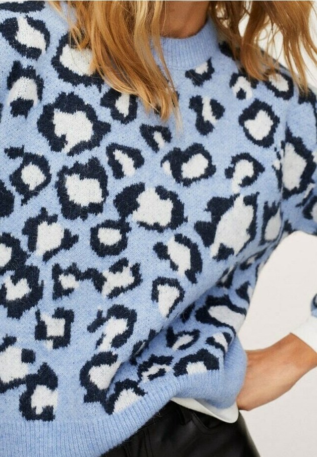 Pull tricoté bleu clair avec motif léopard noir et blanc porté par une personne avec la main sur la hanche et les cheveux blonds ondulés.