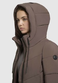 Braune gepolsterte Jacke mit Kapuze, die über eine strukturierte Oberfläche, auffällige Nähte und ein schwarzes, rundes Logo am Ärmel verfügt.