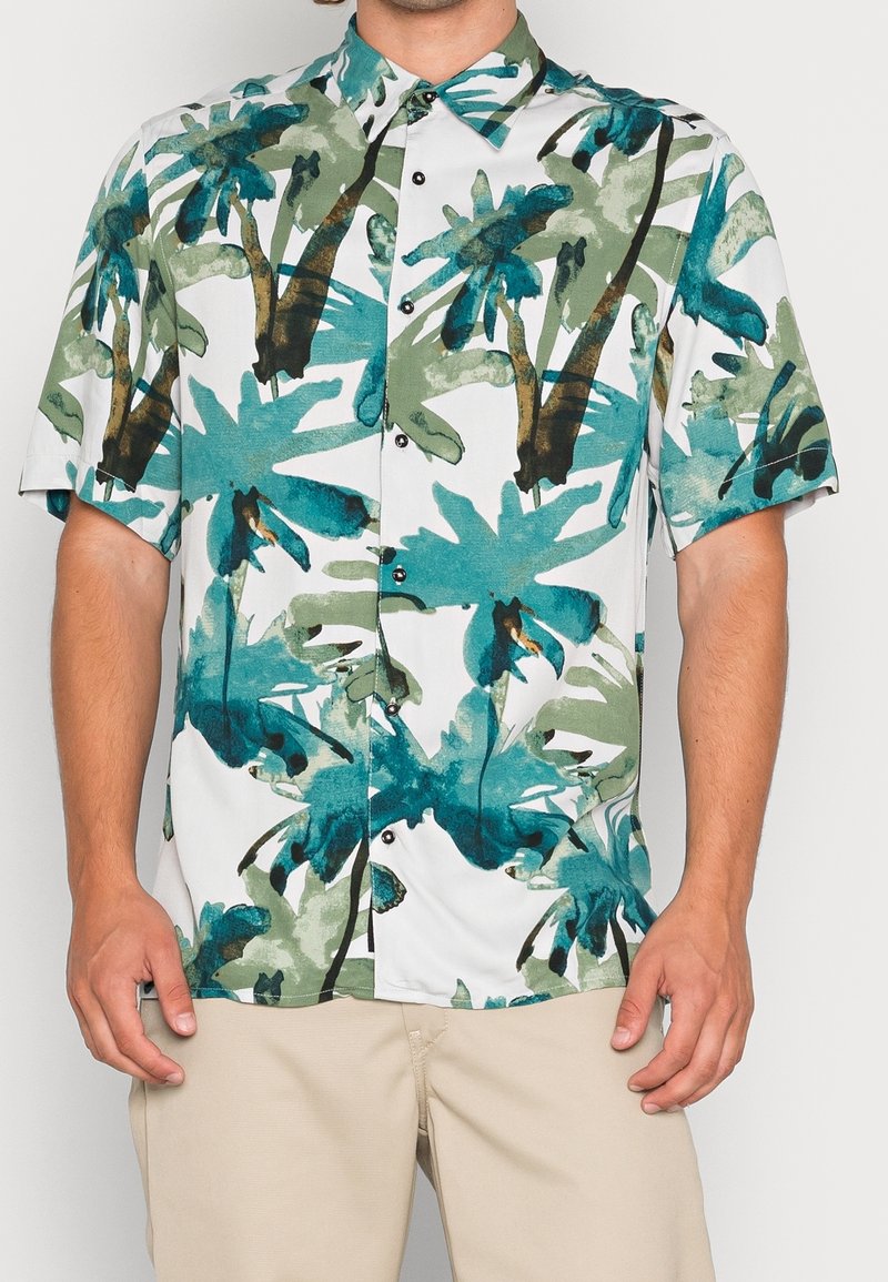 Hombre vestido con camisa de botón de manga corta con estampado de palmeras verdes y azules, combinada con pantalones beige claros, de pie con los brazos relajados.