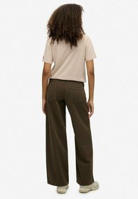 Vrouw met krullend haar, gekleed in een beige T-shirt, donkere wijde broekspijpen en lichte sneakers, staand met de rug naar de kijker op een witte achtergrond.