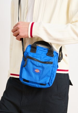 Blått stoff crossover-veske med en glidelås foran lomme og et logo-patch. Har to korte håndtak og en justerbar stropp.