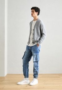 Cárdigan de punto gris claro con botones, combinado con vaqueros cargo azul claro con bolsillos laterales y zapatillas blancas. Textura suave en general.