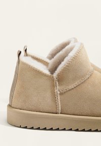 Beżowe zamszowe kapcie wsuwane z puszystym białym futerkiem w środku i teksturowanymi gumowymi podeszwami, pokazane z boku na neutralnym tle.