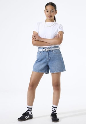 Jonge vrouw met gekruiste armen, gekleed in een wit t-shirt, denim shorts met een witte riem, witte sokken en zwarte sneakers.
