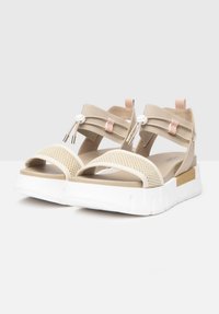 Beige sandals with mesh straps and white platform sole. Elastic bands and gold accents for adjustable fit. Smooth texture, casual design.