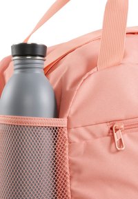 Zaino in tessuto rosa con una tasca laterale in rete che tiene una borraccia in metallo grigio. La zip visibile e i dettagli delle cuciture migliorano la funzionalità.