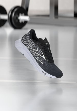 Chaussure de sport noire et grise avec semelle blanche et motif géométrique, flottant au-dessus d'un sol réfléchissant avec équipement de gym flou en arrière-plan.