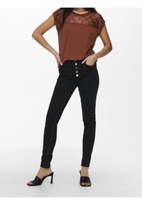 Hnědý top s krajkovými detaily, kombinovaný s černými skinny džíny s vysokým pasem, které mají zlaté knoflíkové akcenty, a černými sandály na podpatku.