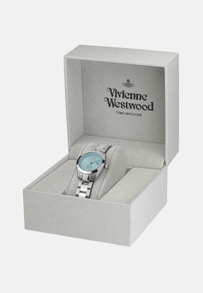 Montre Vivienne Westwood dans une boîte gris clair. Elle présente un bracelet en métal argenté et un cadran rond turquoise avec des index horaires.