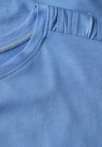 Camiseta azul claro con un escote redondo y detalle fruncido en el hombro. Fabricada en suave algodón con una textura uniforme.