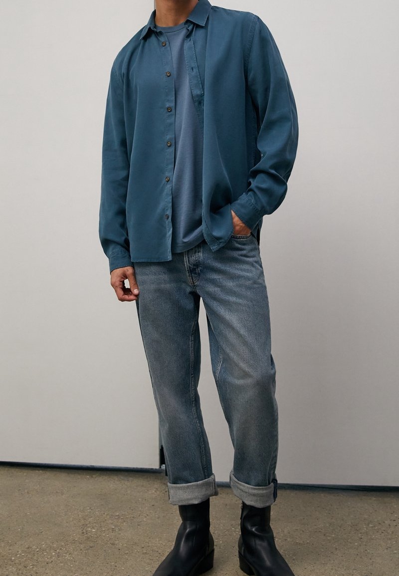 Homme portant une chemise bleue déboutonnée sur un t-shirt bleu, un jean délavé retroussé et des bottines noires, debout devant un fond uni clair.