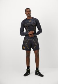 Athletischer Mann steht mit verschränkten Armen, trägt ein langärmliges Kompressionsshirt mit schwarz-gelbem Muster, passende Shorts und schwarze Sneakers mit hohem Schaft.