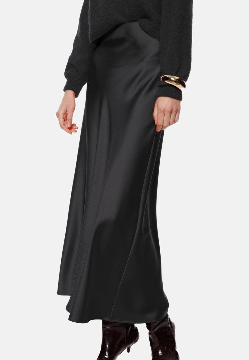 Gonna maxi in satin nero con una texture liscia, caratterizzata da una vita alta e una forma fluida. Messa in risalto da una persona che indossa un maglione scuro e braccialetti.