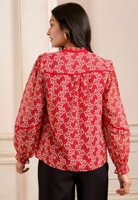 Blusa rossa a fantasia con maniche lunghe, caratterizzata da un design floreale e dettagli arricciati ai polsini. Sul retro presenta una spalla e una piega.