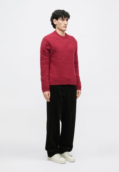 Κόκκινο υφαντό πουλόβερ με υφή, στρογγυλή λαιμόκοψη, ριμπ μανσέτες, συνδυασμένο με μαύρο παντελόνι από χοντρό κοντό ύφασμα. Το μοντέλο φοράει λευκά αθλητικά παπούτσια.
