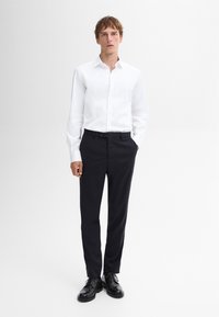 Weißes Button-Down-Hemd mit klassischem Kragen, kombiniert mit dunkelblauen, maßgeschneiderten Hosen und schwarzen Schnürschuhen, vor einem weißen Hintergrund stehend.
