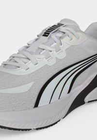 Λευκό και γκρι αθλητικό παπούτσι Puma με μαύρες ρίγες, σχέδιο με κορδόνια και χοντρή μαλακή σόλα, εμφανίζεται σε κοντινή πλάγια όψη.