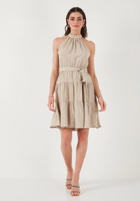 Beige mekko, jossa on halterneck-tyyli, istuva vyötärö, kerroksellinen hame ja sivusolkiyksityiskohta. Kangas on sileä.