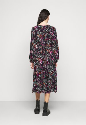 Kobieta z długimi ciemnymi włosami, ubrana w czarną midi sukienkę w kwiaty i czarne botki, stojąca na tle jasnego, jednolitego tła.