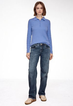 Młoda kobieta stojąca, ubrana w lawendową koszulkę polo z długim rękawem, niebieskie dżinsy i beżowe trampki na jednolitym białym tle.