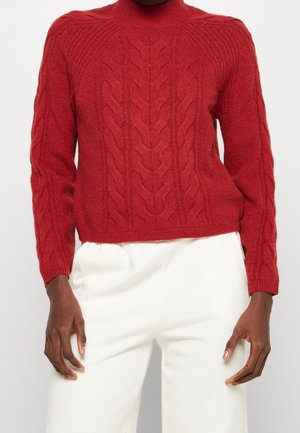 Maglione rosso a trecce con maniche lunghe indossato con pantaloni bianchi a vita alta, mani rilassate ai fianchi su sfondo neutro.