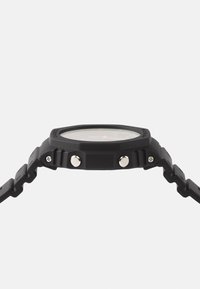 Montre en caoutchouc noir avec un bracelet texturé, de forme ronde, deux boutons-poussoirs argentés et un cadran transparent affichant l'heure avec des accents orange.