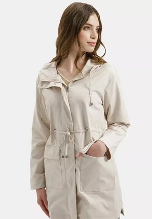 Kobieta w jasnobeżowej kurtce z kapturem, zapinanej na suwak z przodu, z regulacją talii na sznurek i dużymi przednimi kieszeniami, jedna ręka w kieszeni, patrzy w prawo.