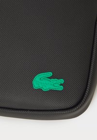 Custodia per laptop nera con texture, caratterizzata da un logo di coccodrillo ricamato verde nell'angolo in basso a sinistra. Cerniera liscia lungo il bordo.