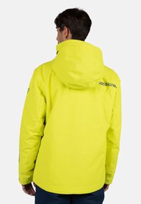 Chaqueta impermeable amarilla con capucha, presenta un logo en la espalda y un acabado texturizado. Las muñecas son ajustables con acentos oscuros.