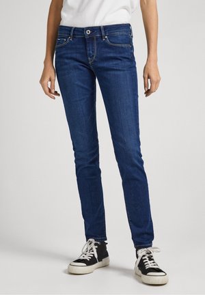 Jean skinny bleu foncé avec un design classique à cinq poches, une légère décoloration et une jambe ajustée, associés à des baskets noires et blanches.