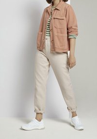 Veste en denim rose clair avec deux poches avant, portée sur une chemise à rayures vertes. Un pantalon beige et des baskets blanches complètent la tenue.