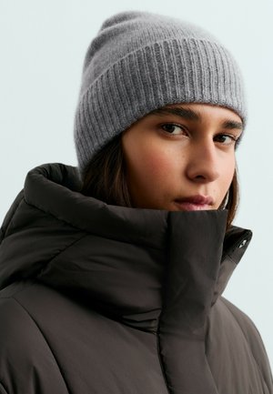 Gros plan d'une personne portant un bonnet tricoté gris et une veste d'hiver noire à col montant, sur un fond clair uni.
