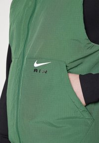 Grön Nike Air-väst med framdragkedja och ficka, hand delvis i fickan, bärs över svart långärmad tröja.