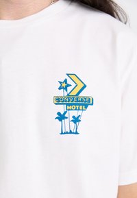 Hvid bomulds T-shirt med et blåt og gult "CONVERSE MOTEL" grafisk design, der inkluderer en stjerne, pil og palme. Korte ærmer.