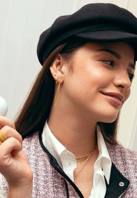 Pendientes de oro con un diseño largo y puntiagudo, y pequeños acentos de aro; collares en capas en tonos rosa y oro; el modelo lleva una gorra negra y una chaqueta de tweed.
