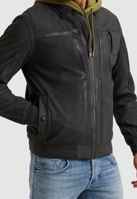 Zwarte leren jasje met een ritssluiting aan de voorkant, geribbelde manchetten en kraag. Beschikt over meerdere zakken en textuurnaden. Gedragen over een groene hoodie.