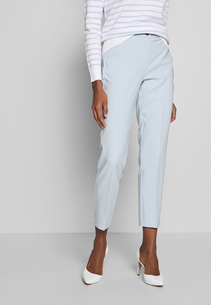 Personne portant un pantalon court sur mesure bleu clair, un pull blanc et gris rayé, et des chaussures blanches à talons hauts, debout sur un sol gris.