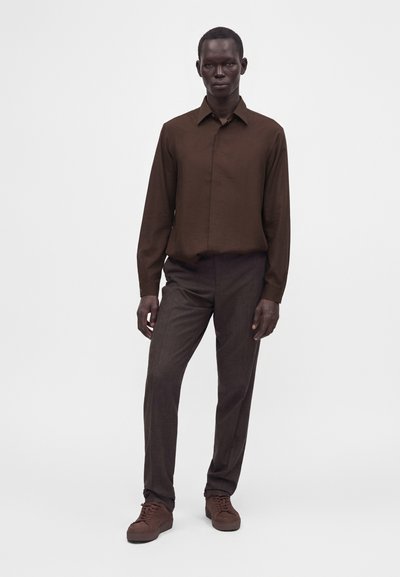 Homme debout portant une chemise marron à manches longues, un pantalon marron foncé et des baskets marron sur un fond clair uni.