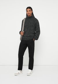 Maglione grigio maculato con collo alto, abbinato a pantaloni cargo neri e sneakers bianche con dettagli beige. Il modello tiene una borsa grigia.