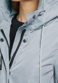 La veste bleu clair est dotée d'une fermeture éclair et à pression, d'une capuche réglable avec cordon de serrage, avec des zips noirs contrastants et des détails à boutons.