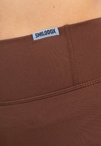 Zbliżenie na brązowy pasek legginsów z widocznym przeszyciem oraz małą szaro-skalową metką z napisem "SMILODOX" na tle jasnej skóry.