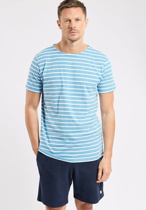 Homme debout portant une chemise à manches courtes rayée bleu clair et blanc et un short bleu foncé sur un fond blanc uni.