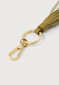 Złoty metalowy breloczek z klamrą, do którego przymocowany jest beżowy skórzany frędzel, na białym tle.