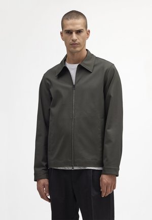 Olivgrön zip-up jacka med spetsig krage, lätt material och två sidofickor. Burit över en ljusgrå skjorta i avslappnad passform.